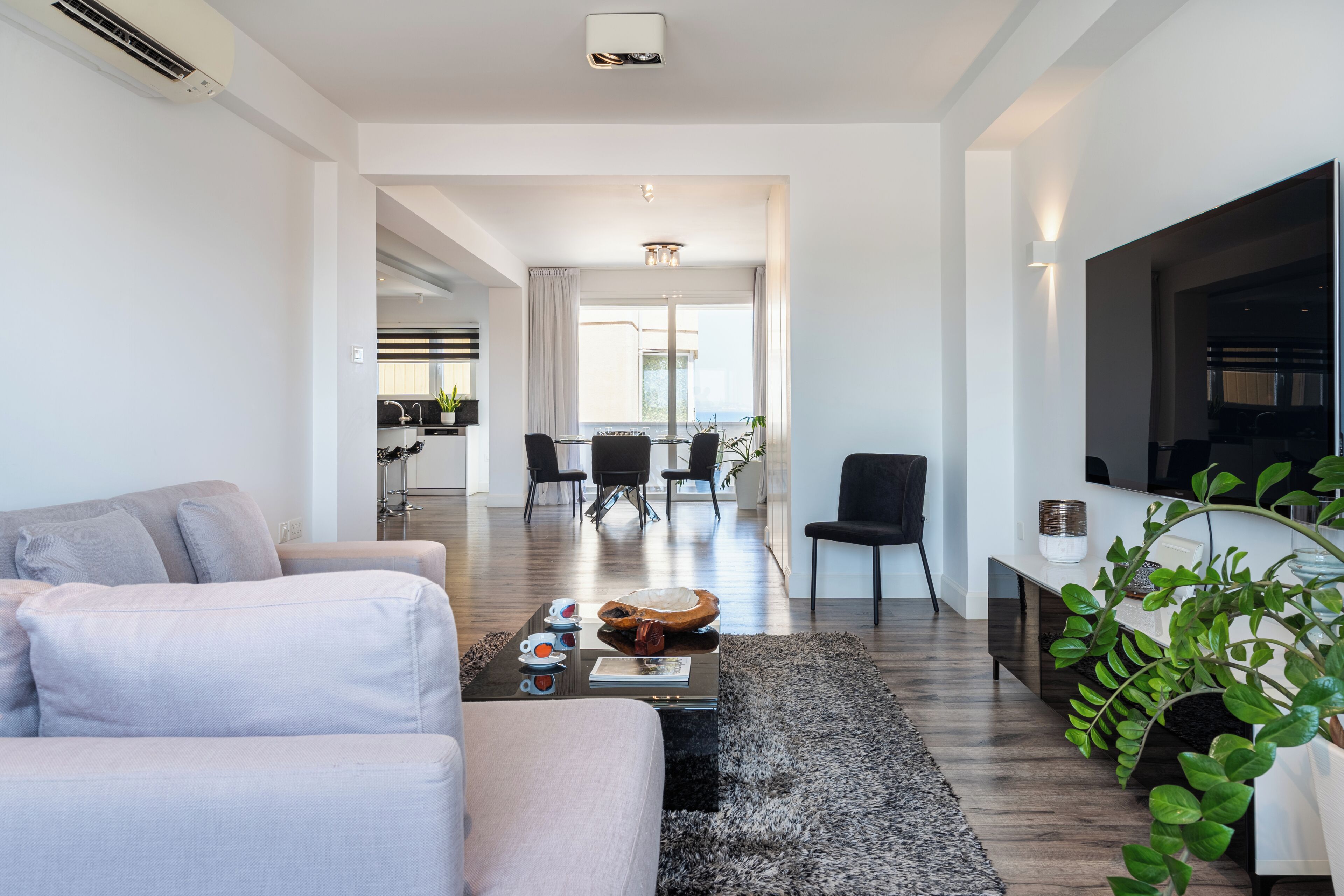 A modern living room with a sofa, a coffee table, and a TV. There's a dining area with chairs and a table in the background.