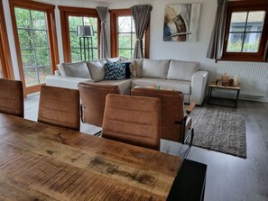 Een moderne woonkamer met een grote houten eettafel, een comfortabele zithoek en uitzicht op groen via grote ramen.