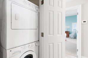 A laundry room with stacked washers and dryers, a white door, and a view into a room with a blue wall and a dresser. - Palms of Dune Allen 304 (Santa Rosa Beach)