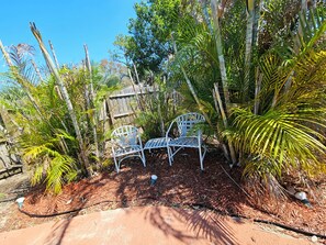Ein Garten mit zwei weißen Metallstühlen, umgeben von verschiedenen tropischen Pflanzen und einem Holzzaun.