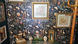 Ein Zimmer mit floraler Tapete, ein grüner Konsolentisch mit Dekoration und ein hölzerner Stand mit einer Laterne.