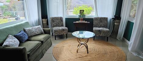 A living room with a green sofa, two patterned armchairs, a glass coffee table, and a woven rug.