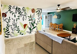 Ein Wohnzimmer mit einer Couch, einem Esstisch, Stühlen, einem Deckenventilator und einer Wand mit einem Blattmuster.