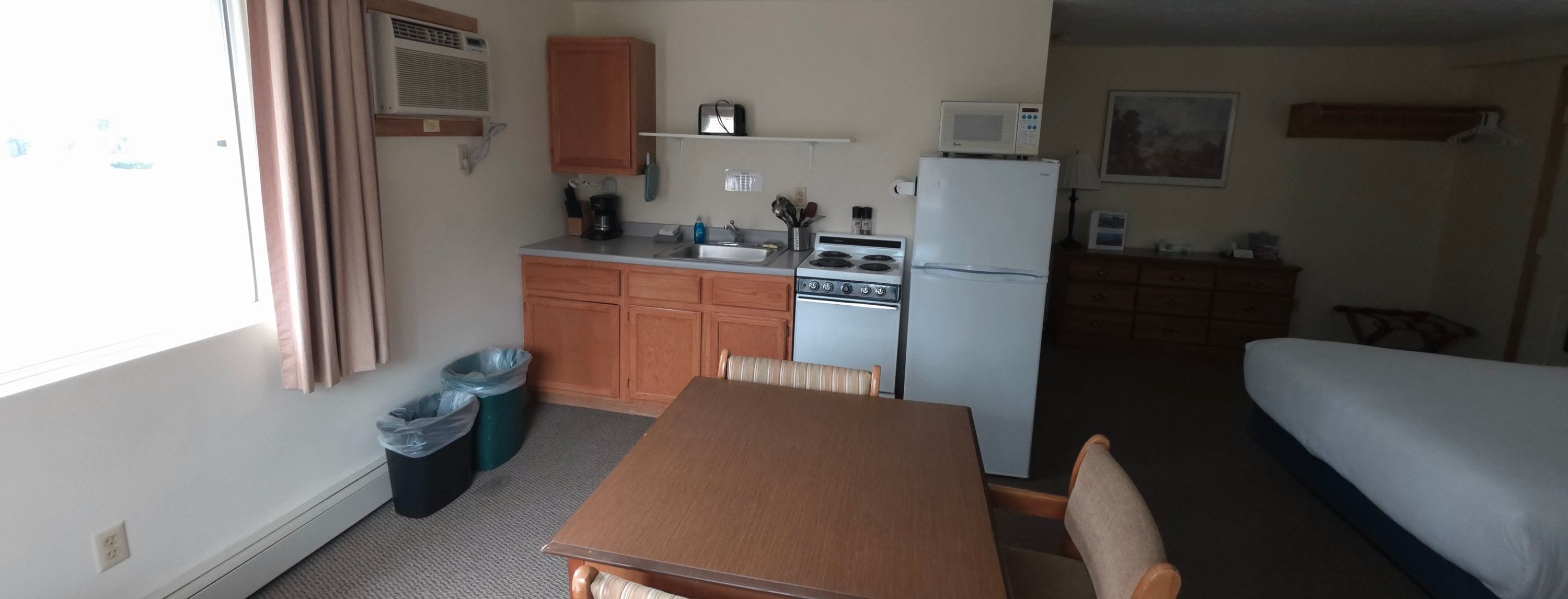 A compact kitchen with a stove, microwave, and refrigerator, a dining table with chairs, and a bed in the adjacent room.