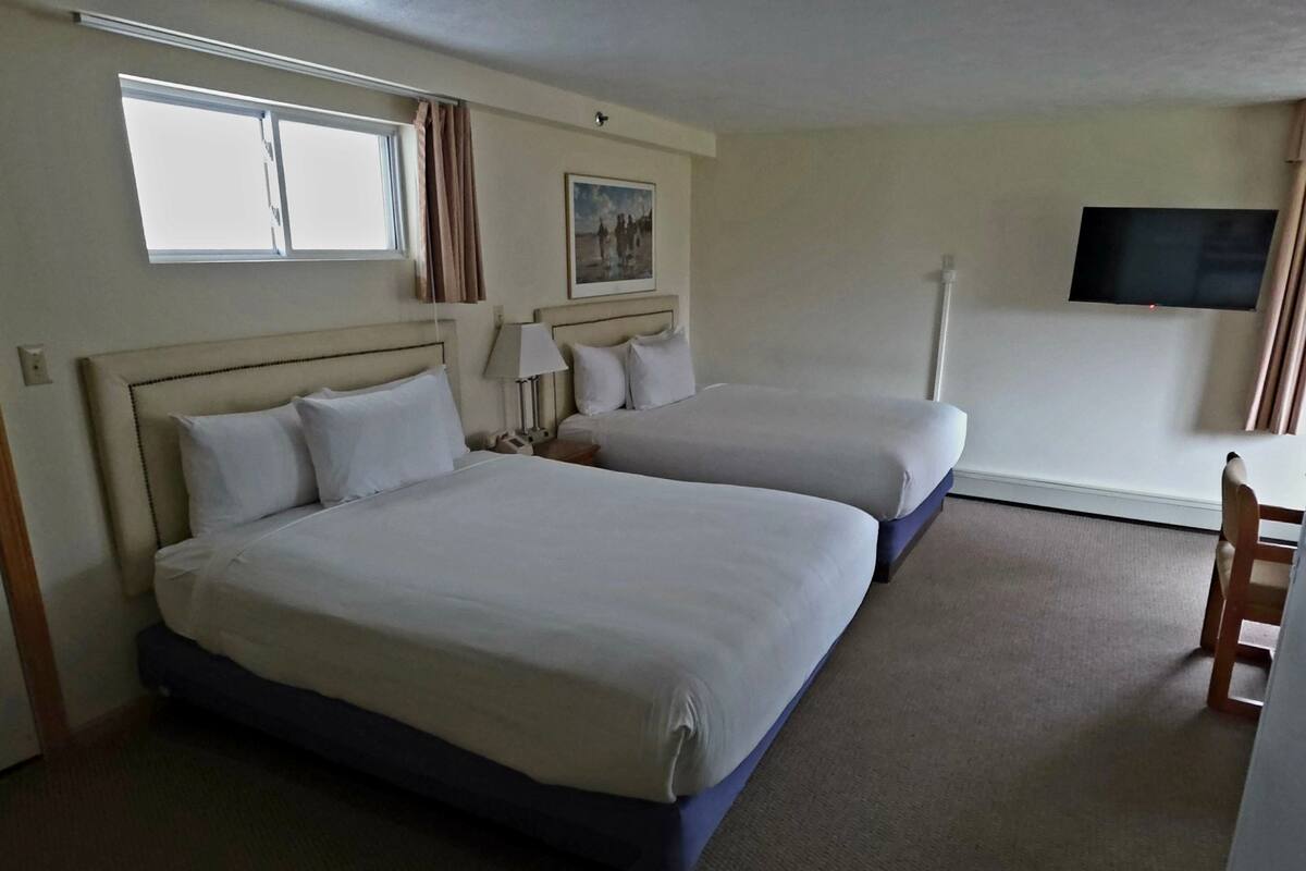 A hotel room with two beds, a television, a window, and a painting on the wall.