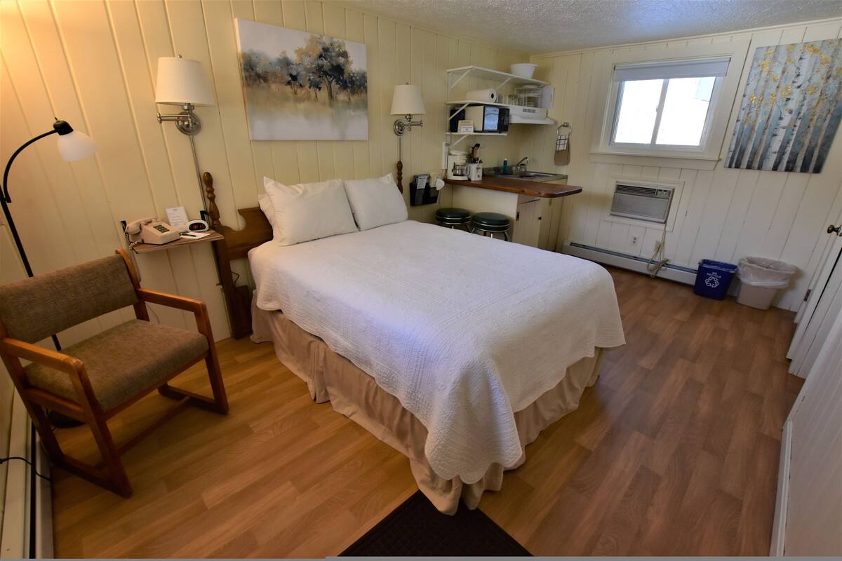 A room with a bed, a chair, a desk with a lamp, a painting, a window, and a kitchenette.