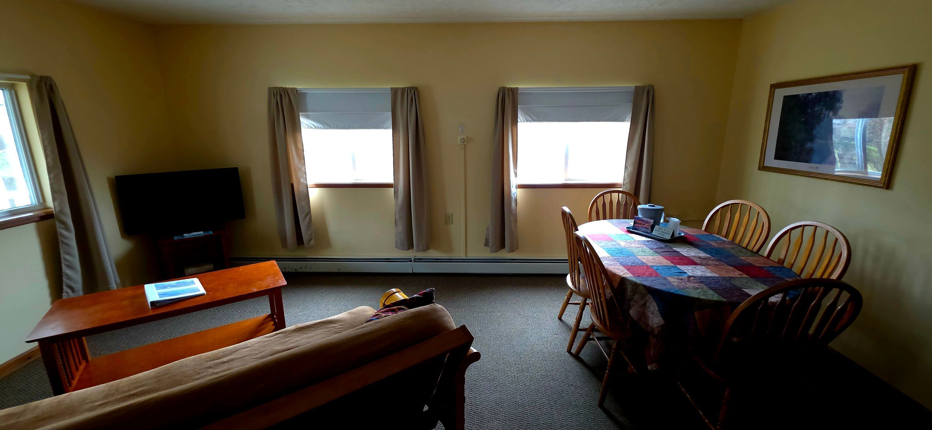 A room with a wooden table, chairs, a bench, a television, and two windows with curtains.