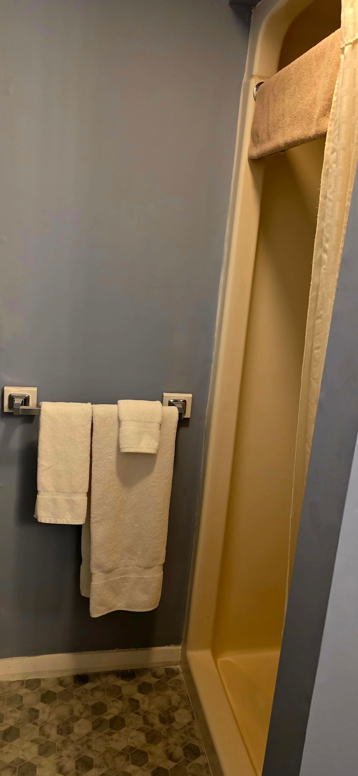 A bathroom with a towel rack holding two towels, a shower with a beige curtain, and a hexagonal tile floor.