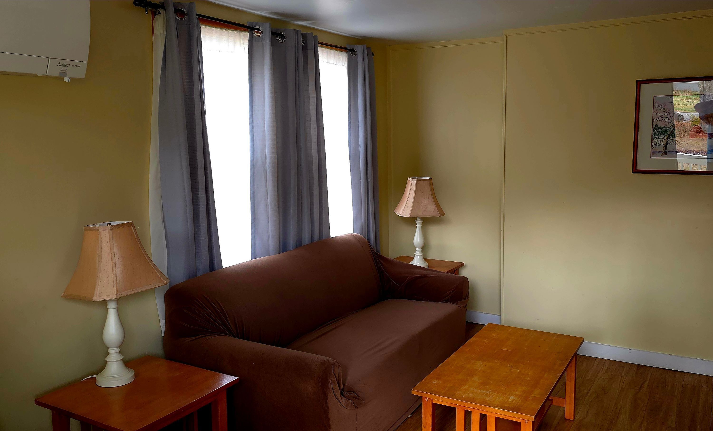 A room with a brown sofa, two wooden side tables, a lamp on each table, a window with curtains, and a framed picture on the wall.