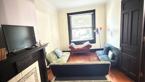 Un salon avec une cheminée, un téléviseur posé sur un meuble, un canapé, une causeuse et une fenêtre avec des stores.