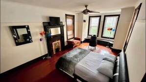 Une chambre à coucher avec un lit, un ventilateur de plafond, une cheminée et une fenêtre avec des stores.