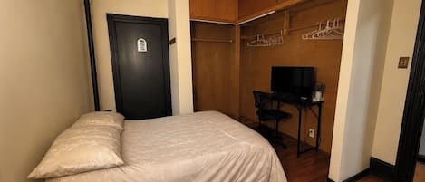 Quarto com cama, armário com roupas penduradas, escrivaninha com cadeira e televisão.
