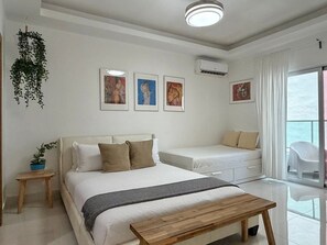Ein modernes Schlafzimmer mit Bett, Nachttisch und kleinem Schreibtisch. An der Wand hängen drei gerahmte Kunstwerke, eine hängende Pflanze und ein Fenster mit Vorhängen.