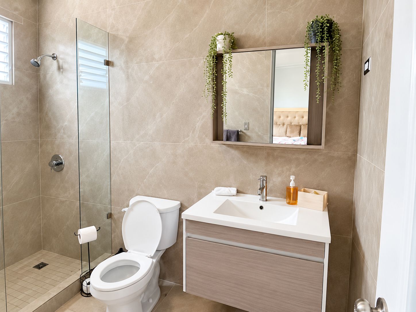 A modern bathroom with a glass shower enclosure, a toilet, a sink with a mirror above, and a plant on the wall.