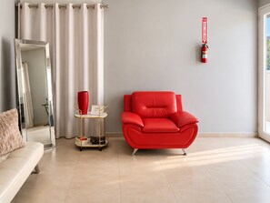 A modern living room with a red leather armchair, a white sofa, a small table with a red vase, and a fire extinguisher mounted on the wall.
