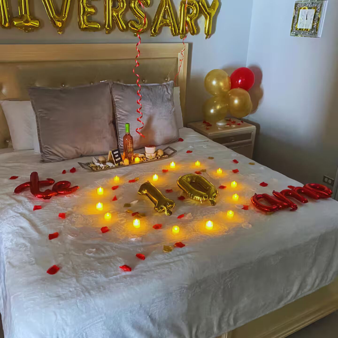 Een bed met een witte spreideken, gouden en rode ballonnen, kaarsen en een fles.