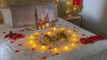 A bed with a white bedspread, gold and red balloons, candles, and a bottle.