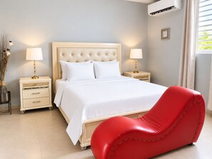 A hotel room with a bed, bedside tables, lamps, a red chair, and an air conditioning unit.