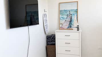 Een kamer met een wandgemonteerde flatscreen-tv, een wit kastje met lades, een rieten mand en een schilderij van een zeilboot in een lijst.