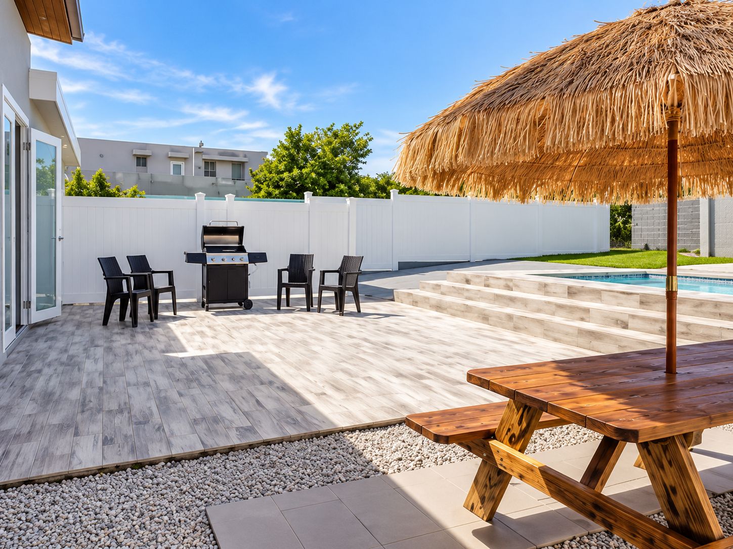 An outdoor patio with a barbecue grill, seating, a wooden table, and a pool in the background.