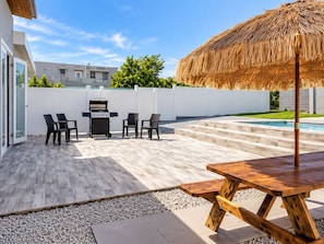 Un patio al aire libre con una parrilla, asientos, una mesa de madera y una piscina de fondo.
