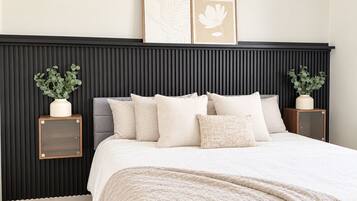 A bedroom with a bed, pillows, a bedside table, a wall-mounted air conditioner, and framed artwork.
