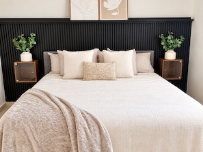 Una cama bien tendida con una manta texturizada, un cabecero oscuro y dos mesitas de noche con plantas en macetas.
