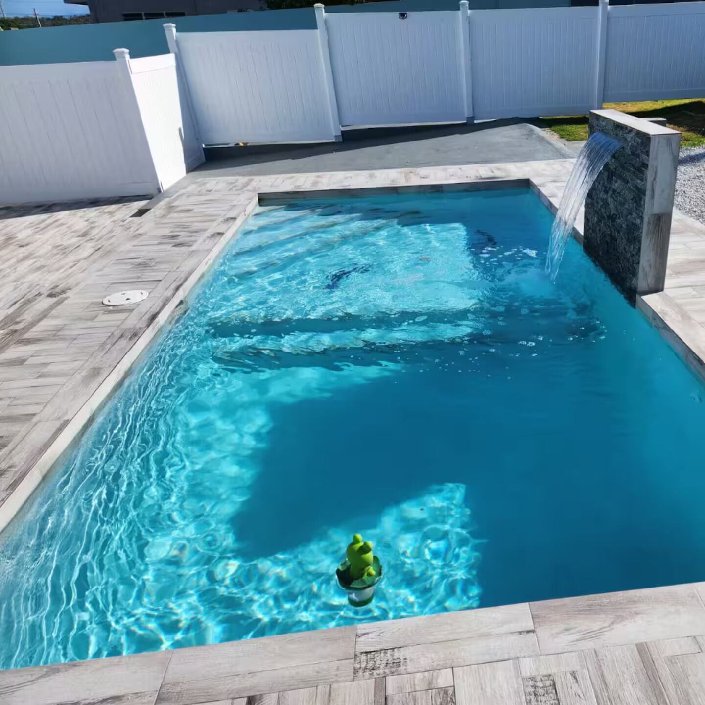A rectangular swimming pool with clear blue water, a small waterfall feature, and a potted plant in the center.