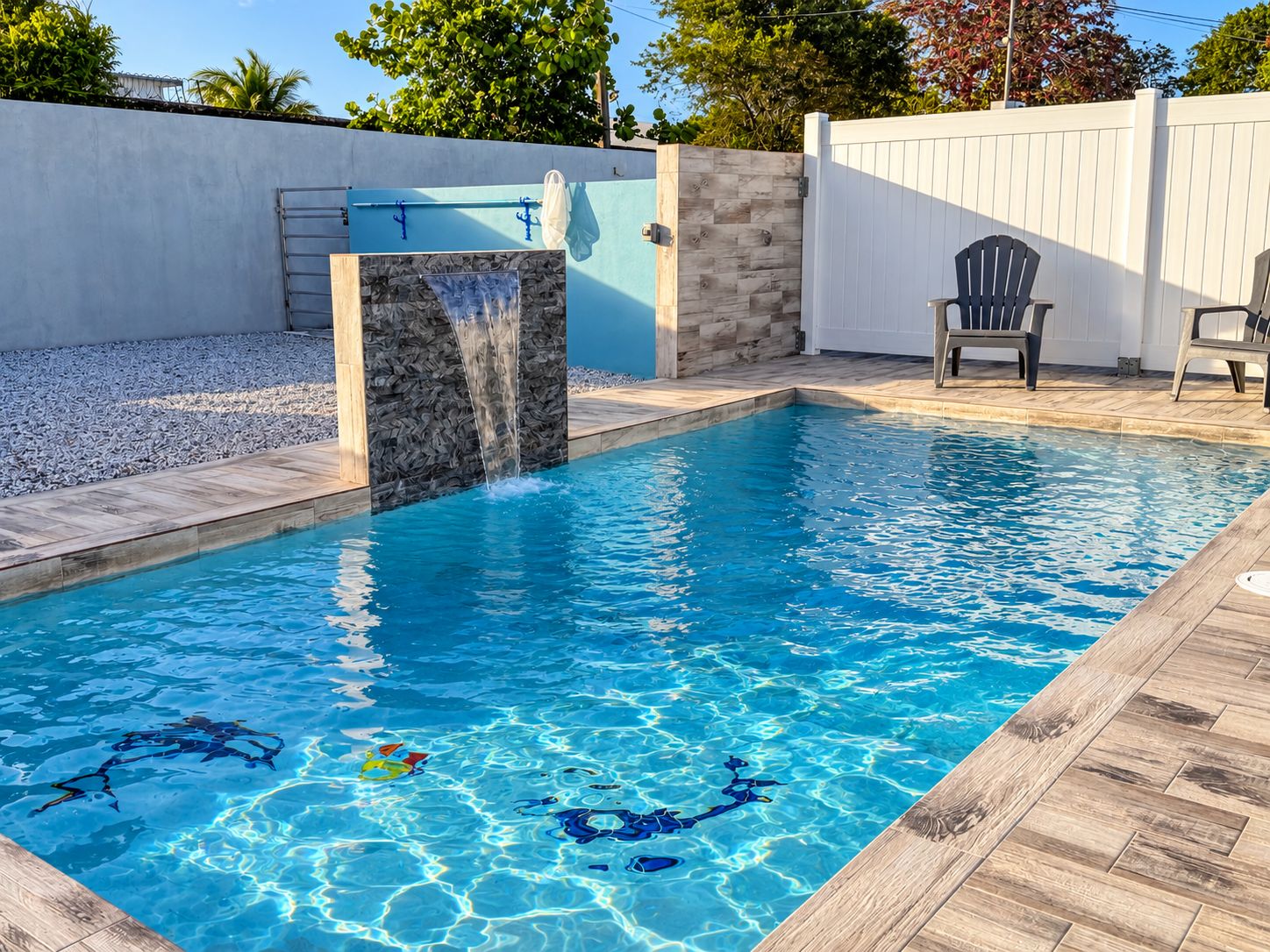 A rectangular swimming pool with a waterfall feature, surrounded by a wooden deck and a white fence.