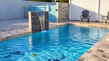 A rectangular swimming pool with a waterfall feature, surrounded by a wooden deck and a white fence.