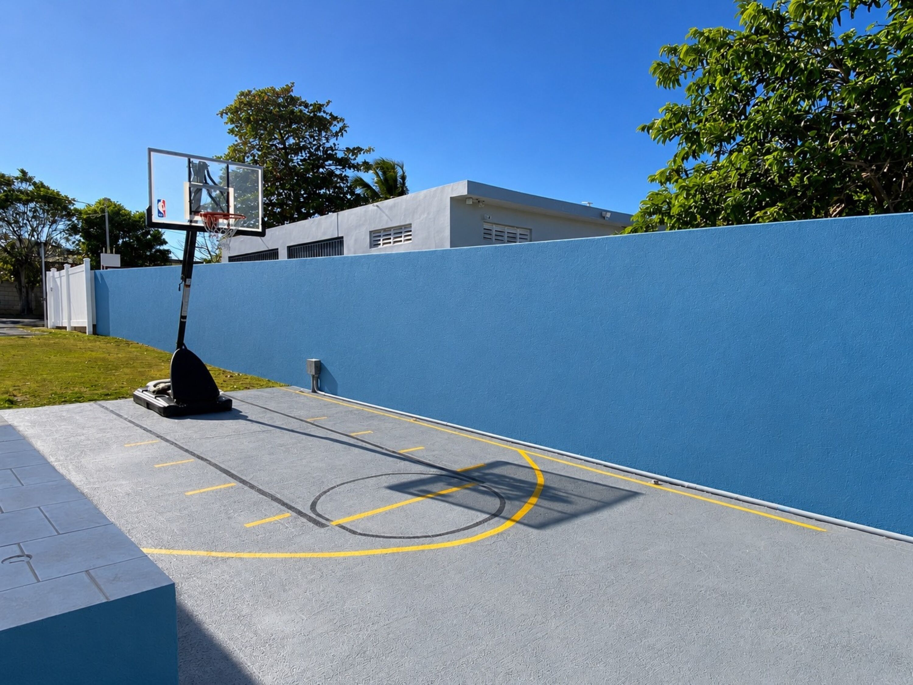 A basketball court with a hoop, a blue wall, and a building in the background.