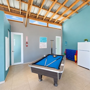 Una mesa de billar en una habitación con paredes color turquesa, techo de madera y un refrigerador blanco.