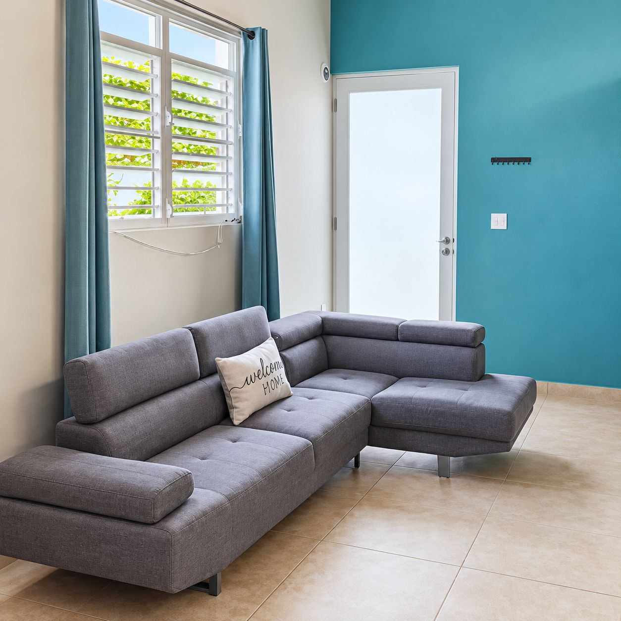 A modern living room with a grey sectional sofa, a teal accent wall, and a window with a view of greenery.