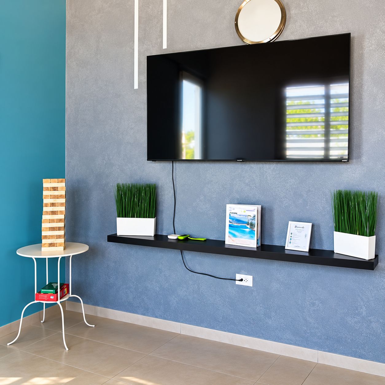 Ein modernes Wohnzimmer mit einem flachbildfernseher an der Wand, einem kleinen runden Tisch mit einer Stapel Bücher, zwei Topfpflanzen und einem gerahmten Zertifikat auf einem schwarzen Regal.