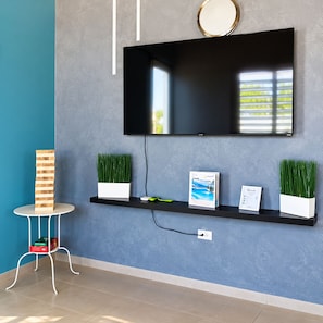 A modern living room with a flat-screen TV mounted on the wall, a small round table with a stack of books, two potted plants, and a framed certificate on a black shelf.