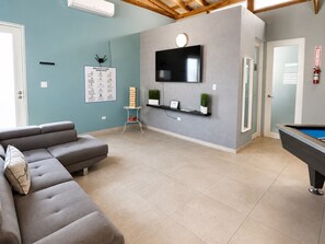 Una sala de estar moderna con un sofá gris, una mesa de billar, un televisor montado y una pequeña mesa con plantas.