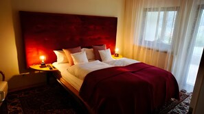 Una habitación de hotel con una cama grande, dos mesitas de noche, una silla y un ventanal con cortinas traslúcidas.