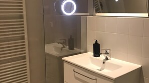 Un baño moderno con cabina de ducha de vidrio, un lavabo blanco con dispensador de jabón negro y un espejo sobre el lavabo.