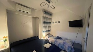 Hotellihuone, jossa on sänky, yöpöytä, lamppu, ilmastointilaite, verhot, seinäkoristeet ja televisio.