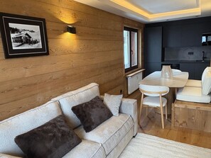 Ein gemütliches Zimmer mit Holzwand, einem gerahmten Bild, einer Couch mit Kissen, einem Esstisch mit Stühlen und einer Küchenzeile.