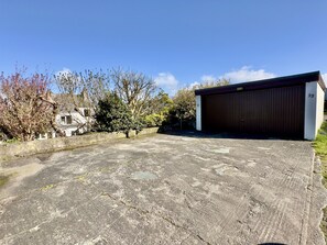 A concrete parking area with a large garage door, surrounded by trees and a white fence. - 22 Treverbyn Road - Spacious Home with amazing estuary views (Padstow)