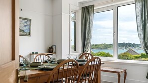 A dining area with a wooden table and chairs, a window with curtains, and a view of the sea. - 22 Treverbyn Road  - Spacious Home with amazing estuary views (Padstow)