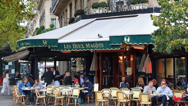 An outdoor café with wicker chairs and tables, a covered seating area, and a sign reading 'LES DEUX MAGOTS'. - DESIGNED BY ACCLAIMED ARCHITECTS IN THE HEART OF SAINT-GERMAIN- RUE DE GRENELLE (Paris)