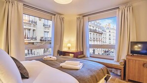 A hotel room with a bed, a TV, a desk with a lamp, and large windows with a view of Parisian buildings. - DESIGNED BY ACCLAIMED ARCHITECTS IN THE HEART OF SAINT-GERMAIN- RUE DE GRENELLE (Paris)