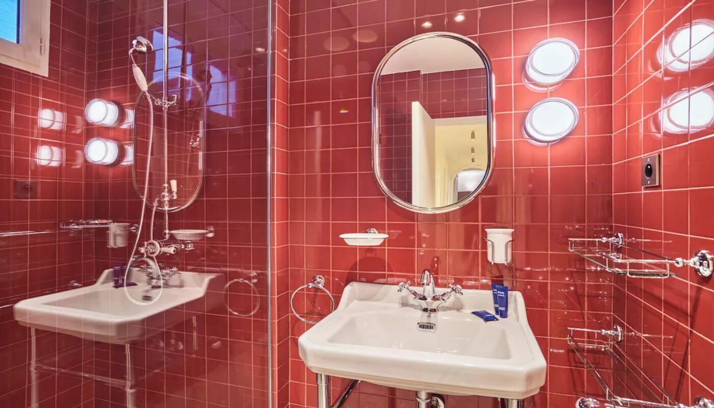 A bathroom with red tiled walls, a large mirror, two sinks, and a shower.