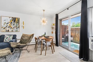 A modern living room with a dining table, chairs, a sofa, and a large window with a view of a wooden deck. - Coastal Comfort Steps From Csu, Pool, Gym, Sauna (Fort Collins)