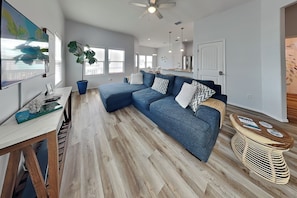 Ein modernes Wohnzimmer mit einem Ecksofa, einem Essbereich mit Tisch und Stühlen, einem großen Fenster und einem Deckenventilator.