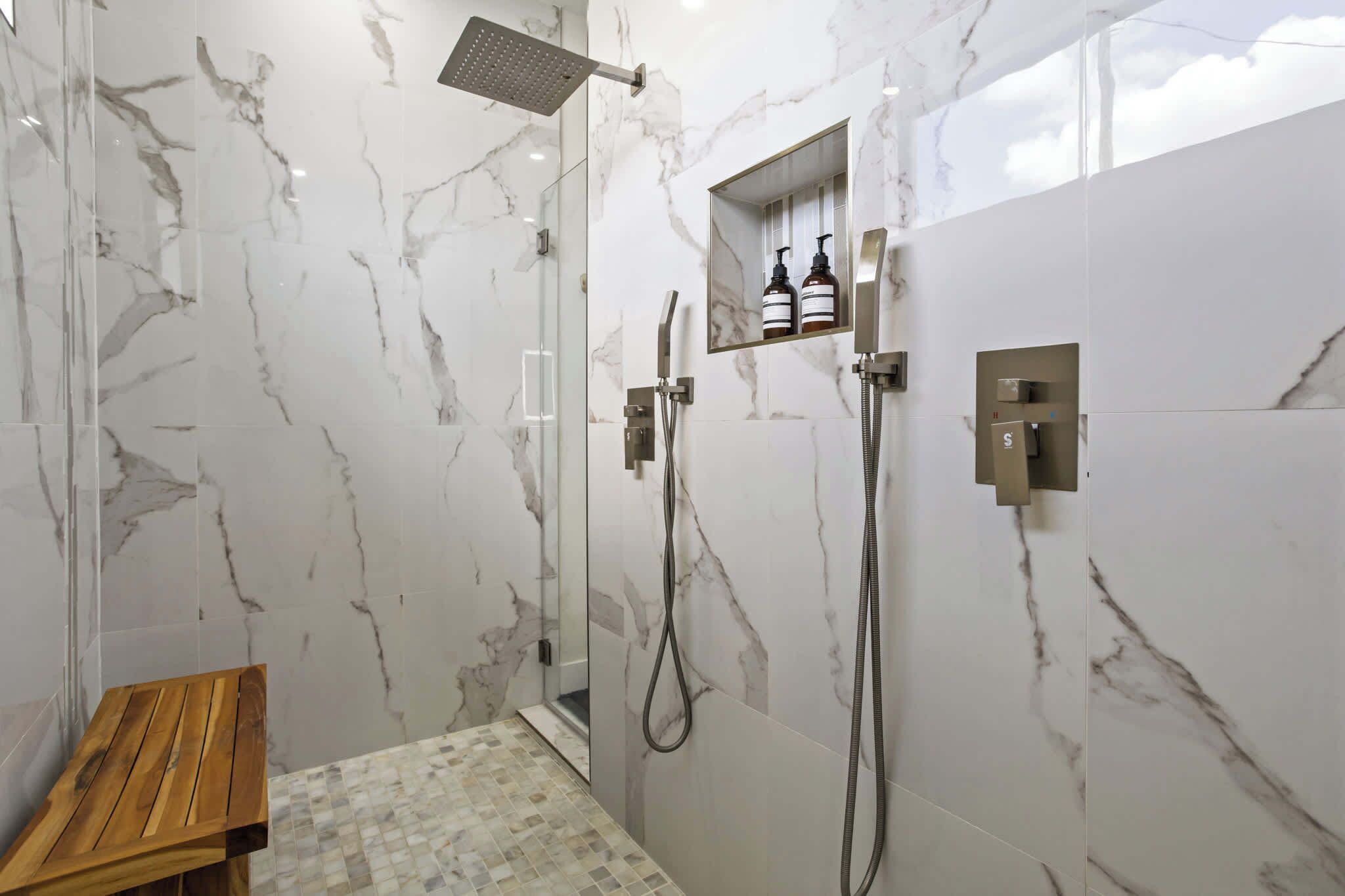 A modern bathroom with a glass-enclosed shower, marble walls, a wooden bench, and a shelf with toiletries.