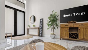 Un salon moderne avec une table basse en bois, une tasse blanche, une pancarte « BIENVENUE », un téléviseur affichant le logo de l’équipe locale et une plante en pot.
