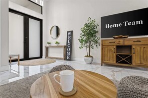 Ein modernes Wohnzimmer mit einem Holztisch, einer weißen Tasse, einem 'WELCOME'-Schild, einem 'Home Team'-Fernseher und einer Topfpflanze.